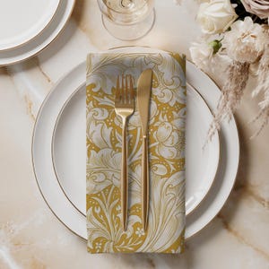 May include: A folded napkin with a floral pattern in gold and white, placed on a white plate with a gold rim. A gold fork and knife are on the napkin. The setting is on a marble surface, with a glass and flowers in the background.