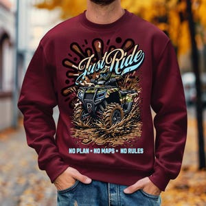 Puede incluir: Sudadera color burdeos con un gr&aacute;fico de una persona conduciendo un ATV por el barro. El gr&aacute;fico incluye el texto "Just Ride" y "NO PLAN - NO MAPS - NO RULES".