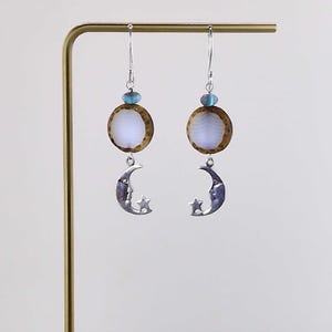 May include: A pair of dangle earrings. Each earring features a silver crescent moon with a face and star, a round, light-colored center, a brown border, and a blue bead at the top. The earrings hang from silver hooks.