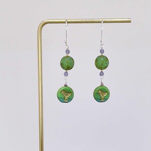 May include: A pair of dangle earrings with a bird design. Each earring features a green, circular bead with a gold bird, a smaller purple bead, and a larger green bead. The earrings hang from silver-colored hooks.