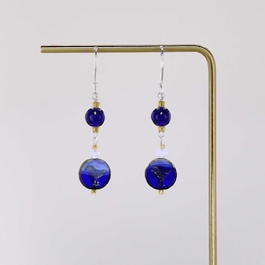 May include: A pair of dangling earrings featuring deep blue glass beads. Each earring has a small, round blue bead at the top, connected to a larger, patterned blue bead below. The earrings hang from silver-colored hooks.