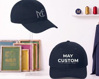 May include: Two navy blue baseball caps. One cap features a silver embroidered logo with the letters MC and a small star. The other cap displays white printed text that reads MAY CUSTOM Print Embroidery Design in a clean, centered font.
