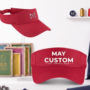 May include: A red visor with a textured fabric finish. One view shows a silver embroidered logo with the letters MC and a star. Another view shows the text MAY CUSTOM Print, Embroidery, Design in white lettering on the front panel of the visor.