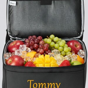 以下が含まれることがあります： 新鮮な果物（リンゴ、ブドウ、イチゴ、マンゴーなど）が詰められた黒い断熱クーラーバッグ。 クーラーには内側にメッシュポケットがあり、前面には金色の文字で「Tommy」と書かれています。