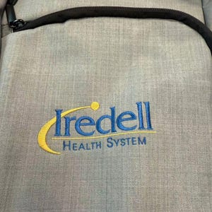 Puede incluir: Una mochila gris con cremallera y ribete negros. La mochila presenta el texto bordado "Iredell Health System" en azul y amarillo.