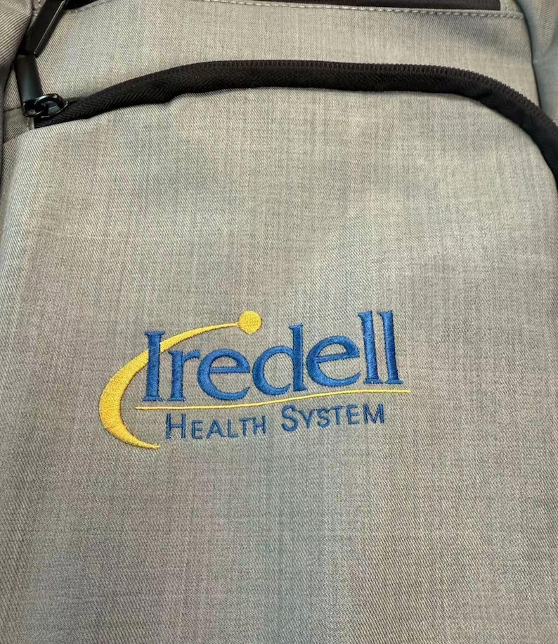 Puede incluir: Una mochila gris con cremallera y ribete negros. La mochila presenta el texto bordado "Iredell Health System" en azul y amarillo.