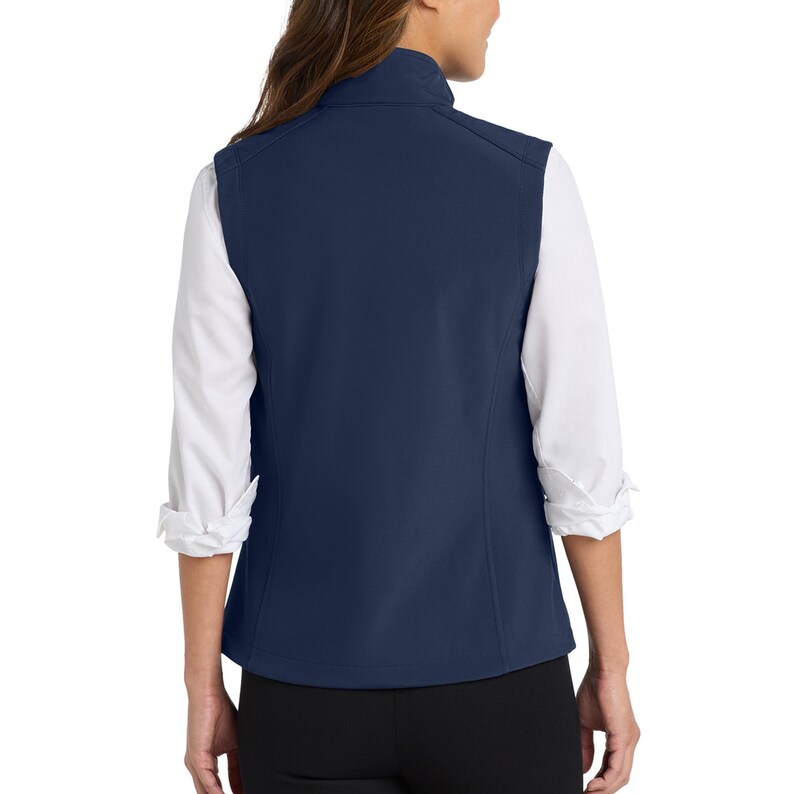 May include: A solid navy blue sleeveless vest featuring a high collar and a smooth, matte finish. The vest is worn over a white long-sleeved shirt with cuffed sleeves and is paired with black trousers, showing the back view of the garment.