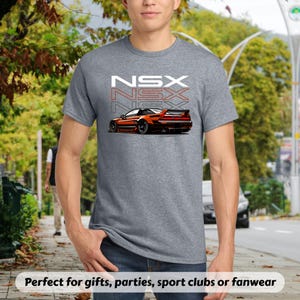 May include: A gray t-shirt featuring a stylized orange sports car graphic with the word "NSX" stacked three times in white and red. The shirt is made of a soft material and is suitable for casual wear.