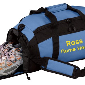 May include: A blue and black duffel bag with a dedicated side shoe compartment containing a pair of grey and orange athletic sneakers. The bag features black webbing straps, a padded shoulder strap, and the yellow text Ross Name Here.