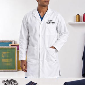 May include: A white lab coat featuring a notched lapel, long sleeves, and a button-down front. The chest pocket displays the black text MAY CUSTOM. Two large patch pockets are located at the waist, and a blue shirt is visible underneath the collar.