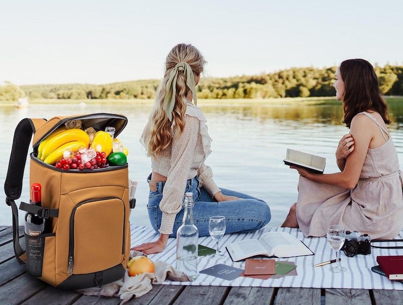 Puede incluir: Una nevera tipo mochila aislante de color marr&oacute;n llena de fruta, incluyendo pl&aacute;tanos y uvas, y una botella de vino. La mochila tiene correas negras y un bolsillo lateral. Dos personas est&aacute;n sentadas cerca del agua con una manta de picnic, libros y bebidas.