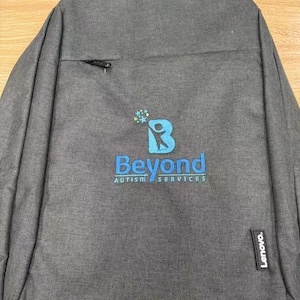 A dark grey backpack features an embroidered logo for Beyond Autism Services in blue and teal on the front pocket.
