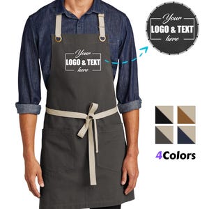 May include: A charcoal gray apron with tan straps and a waist tie. The apron has two front pockets and features the text "Your LOGO & TEXT here". A circular design with the same text is shown, along with color options.