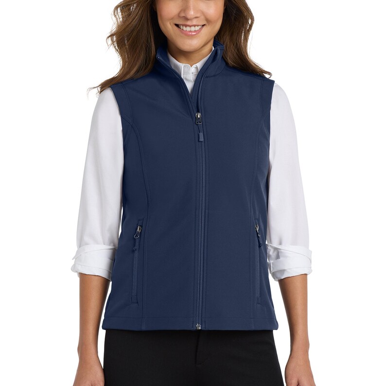 May include: A navy blue sleeveless waistcoat featuring a full-length front zip, a stand-up collar, and two side pockets with zip closures. The waistcoat is worn over a white long-sleeved collared shirt with the sleeves rolled up to the elbows.