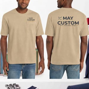 May include: A tan short-sleeve t-shirt with a chest pocket. The front features a small logo with the text MAY CUSTOM. The back displays the text MAY CUSTOM Print. Embroidery. Design in dark lettering. The shirt is paired with blue denim jeans.