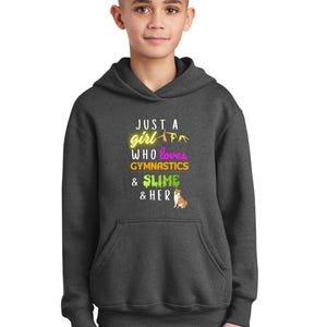 Puede incluir: Una sudadera con capucha gris oscuro con el texto "Just a girl who loves gymnastics & slime & her" en amarillo, rosa y verde. Un peque&ntilde;o perro de dibujos animados est&aacute; en la esquina inferior derecha.
