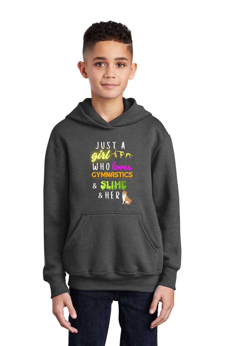 Puede incluir: Una sudadera con capucha gris oscuro con el texto "Just a girl who loves gymnastics & slime & her" en amarillo, rosa y verde. Un peque&ntilde;o perro de dibujos animados est&aacute; en la esquina inferior derecha.