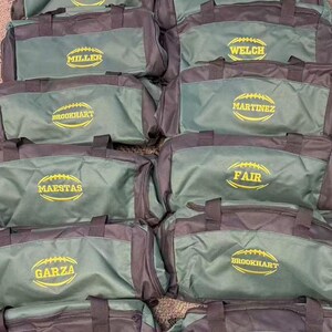 May include: A collection of dark green and black duffel bags, each featuring a yellow embroidered football graphic with a unique name stitched in the center, including Miller, Welch, Brookhart, Martinez, Maestas, Fair, and Garza.