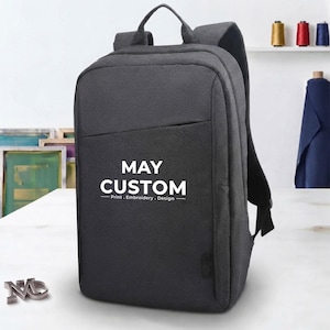 A charcoal grey custom backpack stands on a white table with the text MAY CUSTOM printed in white on the front pocket.