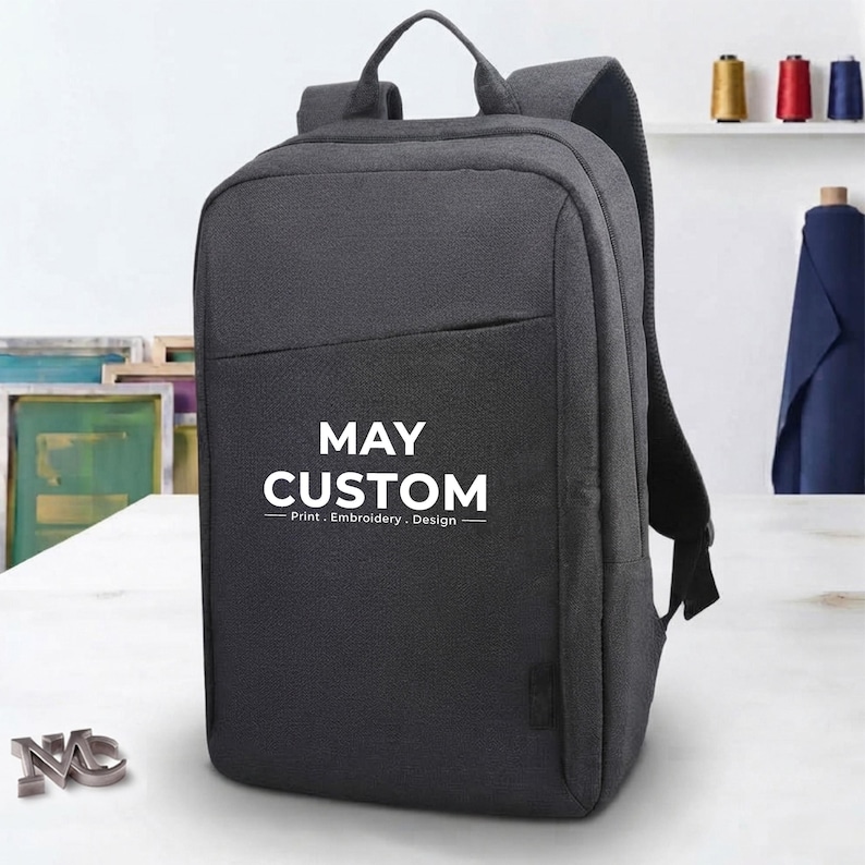 A charcoal grey custom backpack stands on a white table with the text MAY CUSTOM printed in white on the front pocket.