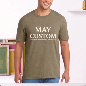 May include: An olive green short-sleeve t-shirt featuring the cream-colored text MAY CUSTOM centered on the chest, with the smaller text Print . Embroidery . Design underneath, flanked by two horizontal lines in a matching cream color.