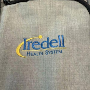 K&ouml;nnte beinhalten: Ein grauer Rucksack mit schwarzem Rei&szlig;verschluss und schwarzem Riemen. Der Rucksack hat den gestickten Text "Iredell HEALTH SYSTEM" in Blau und Gelb.