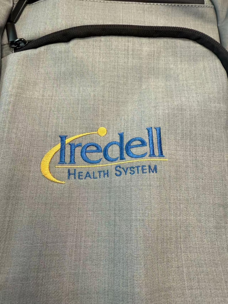 K&ouml;nnte beinhalten: Ein grauer Rucksack mit schwarzem Rei&szlig;verschluss und schwarzem Riemen. Der Rucksack hat den gestickten Text "Iredell HEALTH SYSTEM" in Blau und Gelb.