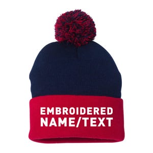 May include: A navy blue and red knit beanie with a burgundy pom-pom. The red cuff features the white text "EMBROIDERED NAME/TEXT". This winter hat is a customizable accessory.