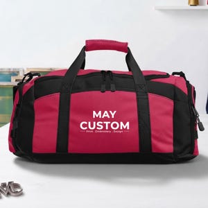 May include: A pink and black duffel bag featuring a top handle and black zipper closures. The front of the bag displays the white text MAY CUSTOM Print . Embroidery . Design. The bag has black side panels and black straps extending from the base.