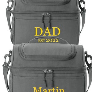 以下が含まれることがあります： 2つのグレーの断熱ランチバッグ。 上部のバッグには、黄色で「DAD」の文字と下に「EST 2022」があります。 下部のバッグには、黄色で「Martin」という名前があります。 どちらのバッグにも上部のハンドル、ショルダーストラップ、ジッパー開閉部があります。