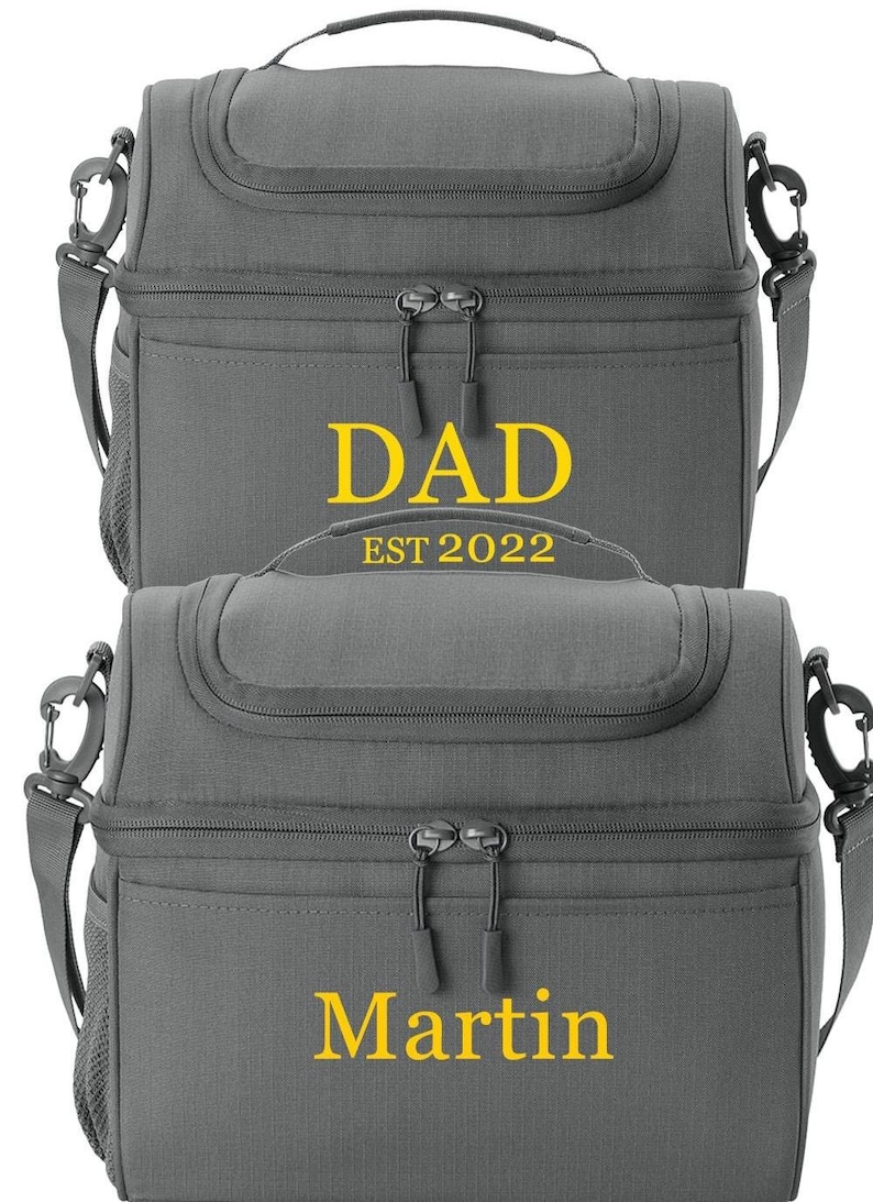 以下が含まれることがあります： 2つのグレーの断熱ランチバッグ。 上部のバッグには、黄色で「DAD」の文字と下に「EST 2022」があります。 下部のバッグには、黄色で「Martin」という名前があります。 どちらのバッグにも上部のハンドル、ショルダーストラップ、ジッパー開閉部があります。
