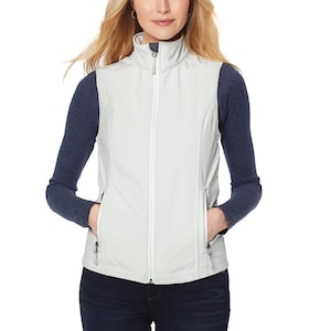 May include: A light grey softshell vest featuring a high collar, a full-length white front zip, and two side pockets with dark grey zip pulls. The vest is worn over a long-sleeved navy blue shirt and dark blue denim jeans.