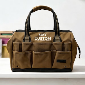 May include: A brown tool bag with black trim and handles, featuring multiple exterior pockets and a silver printed logo that reads MAY CUSTOM Print. Embroidery. Design. The bag has a reinforced base and metal grommets on the front lower corners.