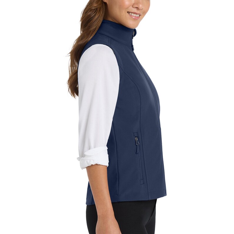 May include: A navy blue sleeveless vest featuring a high collar and a full-length front zip. The vest includes side pockets with vertical zip closures and is worn over a white long-sleeved shirt with rolled-up cuffs and black trousers.