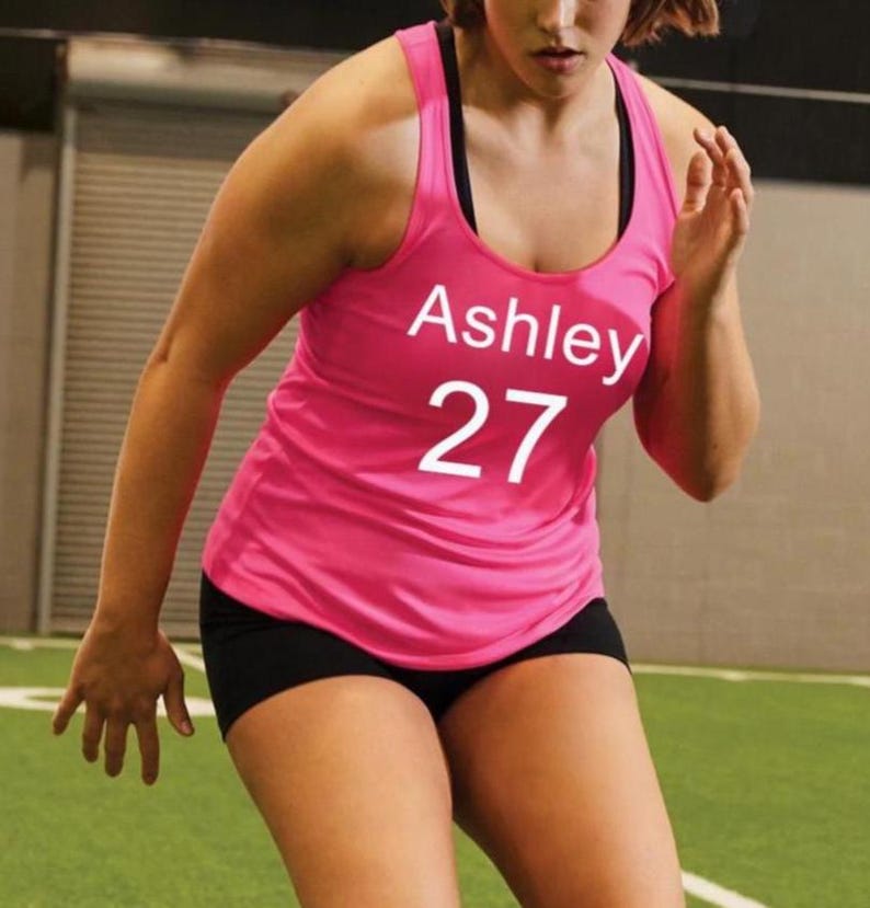 May include: A pink athletic tank top with the name "Ashley" and the number "27" printed in white. The tank top is paired with black athletic shorts. The person is running on a green field.