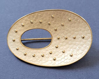 Könnte beinhalten: Eine ovale Brosche aus Silber mit weißer Emaille-Oberfläche. Die Brosche hat ein sternförmiges Muster und ein kleines Loch in der Mitte.