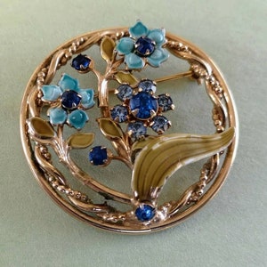 May include: A vintage gold-tone floral brooch. The brooch has a circular design with a detailed border. It features blue and turquoise enamel flowers with dark blue rhinestone centres and green enamel leaves. A classic piece of jewellery.