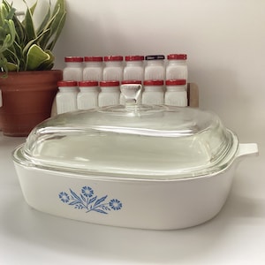 May include: White casserole dish with a blue floral design and a clear glass lid. The dish is sitting on a white surface.