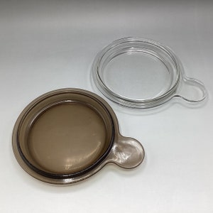 May include: A brown glass lid with a handle and a clear glass lid with a handle. Both lids are round and have a raised edge.