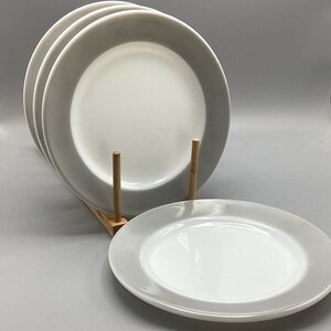 May include: Set of four white ceramic dinner plates with a gray rim. The plates are stacked on a wooden stand.