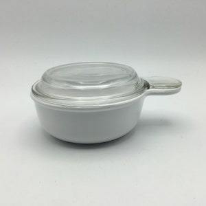 May include: White ceramic casserole dish with a clear glass lid and a handle. The dish is round and has a smooth surface.