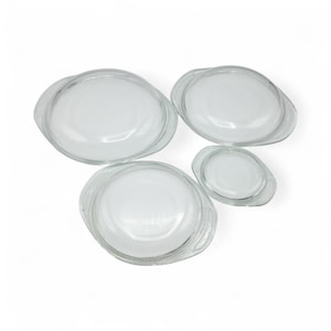 May include: Four clear glass casserole dish lids of varying sizes are arranged on a white background. Each lid has a rounded oval shape with a slightly raised rim and two small handles on either side. The glass is transparent.