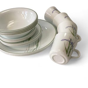 May include: A set of white ceramic bowls and plates with a green and purple floral design. The bowls are stacked and the plates are arranged in a circle around them. There are also four white ceramic mugs with the same floral design.