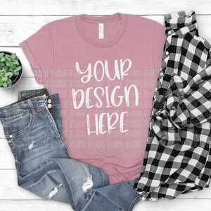 May include: A dusty rose t-shirt with the text "YOUR DESIGN HERE" in white script, paired with distressed blue jeans and a black and white plaid shirt. A small potted succulent sits nearby.