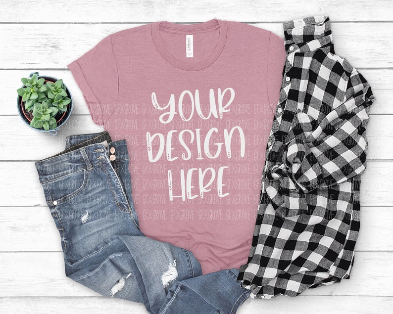 May include: A dusty rose t-shirt with the text "YOUR DESIGN HERE" in white script, paired with distressed blue jeans and a black and white plaid shirt. A small potted succulent sits nearby.