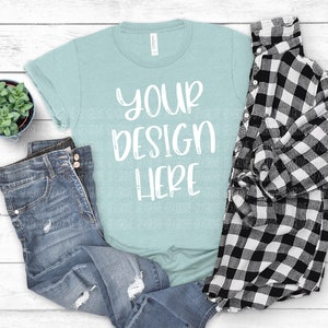 May include: A light blue t-shirt with the text "YOUR DESIGN HERE" in white. The t-shirt is laid out on a white wooden surface with a pair of blue jeans and a black and white plaid shirt.