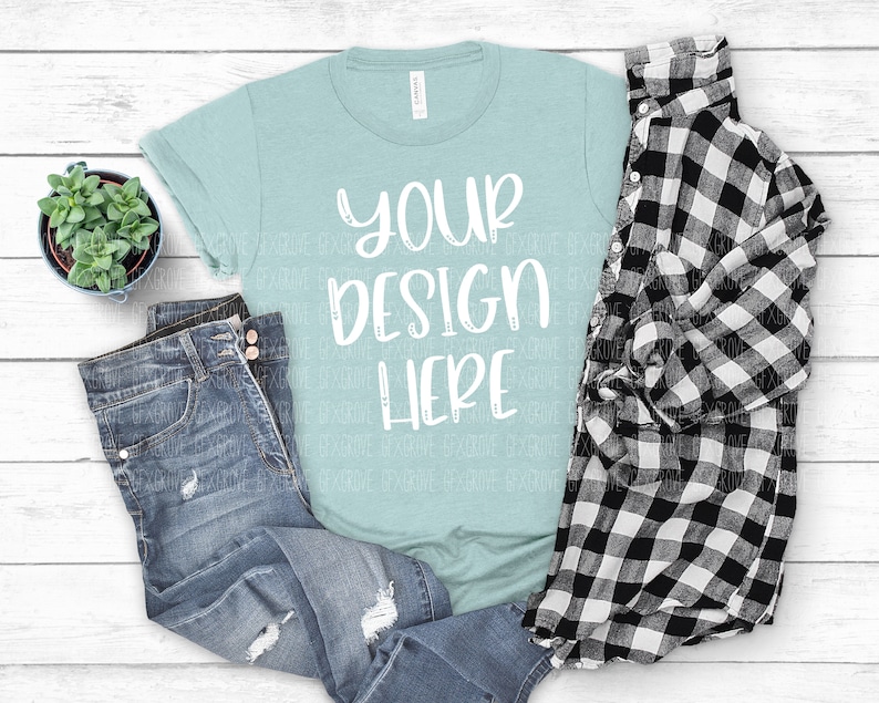 May include: A light blue t-shirt with the text "YOUR DESIGN HERE" in white. The t-shirt is laid out on a white wooden surface with a pair of blue jeans and a black and white plaid shirt.