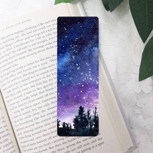 Puede incluir: Un marcapáginas de acuarela con un cielo nocturno azul oscuro y morado con estrellas blancas. El cielo está pintado con una lavada de acuarela y las estrellas están dispersas por el cielo. Hay árboles verde oscuro en primer plano.