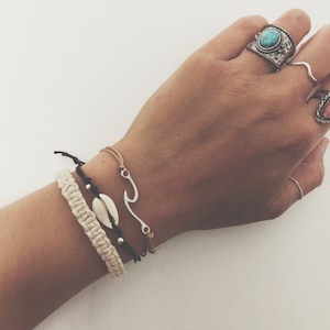 Könnte beinhalten: Ein Handgelenk mit vier Armbändern. Die Armbänder bestehen aus schwarzem Band, braunem Band, weißem Band und einem silbernen Anhänger in Form einer Welle. Der Anhänger ist an einem braunen Bandarmband befestigt.