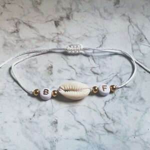 Könnte beinhalten: Weißes Armband aus Schnur mit goldenen Perlen und weißen Perlen mit den Buchstaben "B" und "F". Ein weißer Muschelanhänger befindet sich in der Mitte des Armbands.